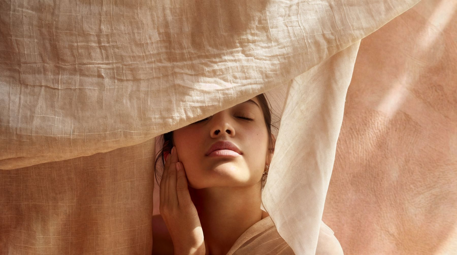 A woman with her eyes closed under soft draped linen, warm light on her skin
