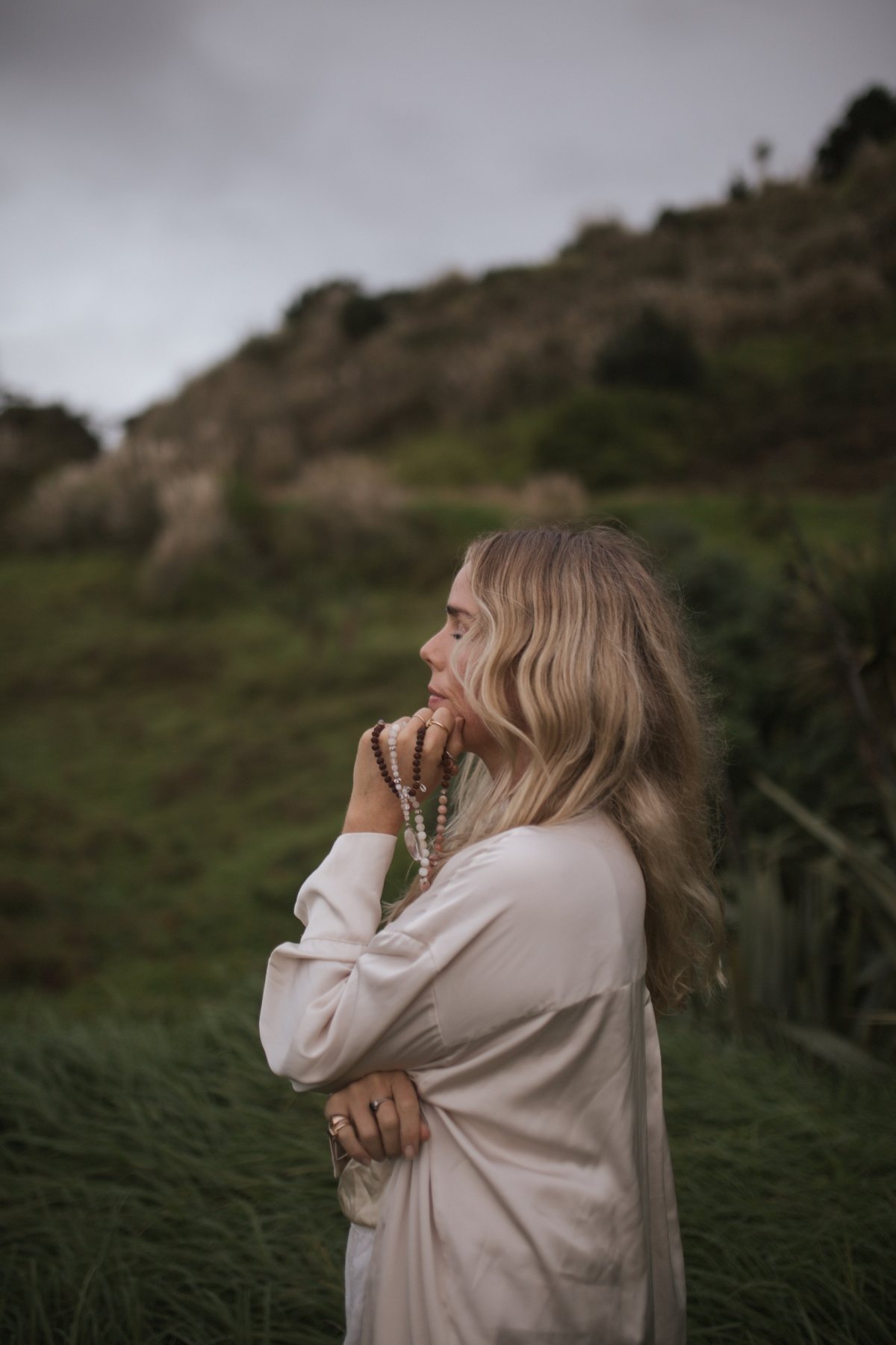 Lauren in a silk shirt, holding mala beads, on a green Mangawhai hillside